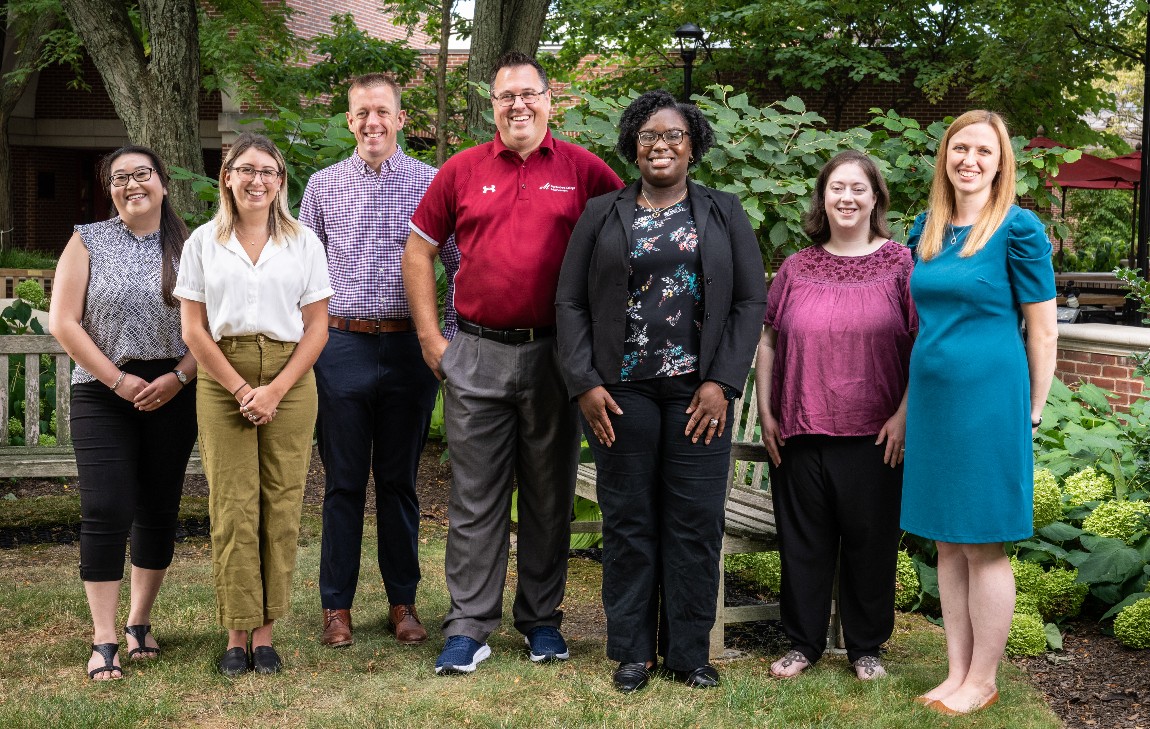 A group of seven people stand in a row outside, smiling for the camera.